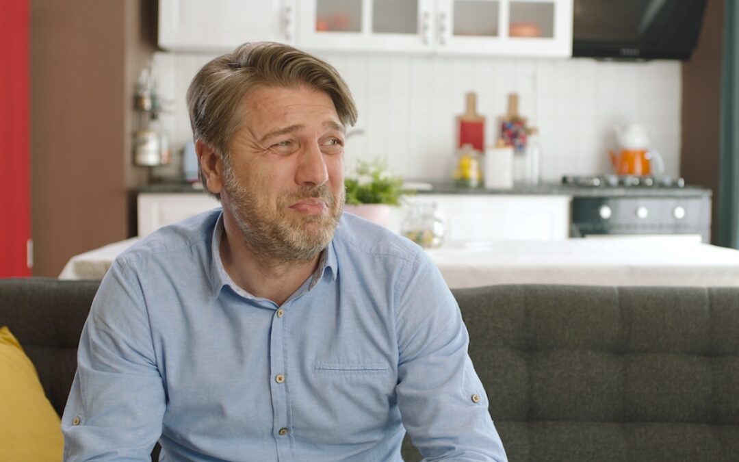 A middle-aged man in a blue shirt sits in a livingroom. He is making a disgusted face, trying to determine the source of the bad smell coming from his heating system.