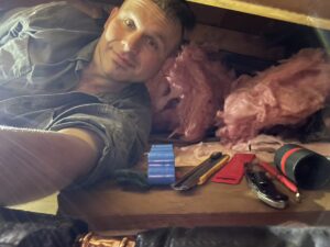 A local Worcester County HVAC technician takes a selfie from within a tight crawlspace, surrounded by insulation, exposed beams, and ductwork.
