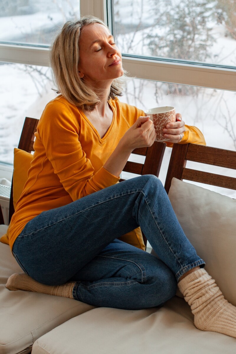 A woman in a yellow sweater is sitting by a window holding a mug of tea with both hands. Outside we can see it is winter, but she is perfectly warm and cozy thanks to her heat pump installed by Turkey Heating and Cooling in Central Massachusetts.